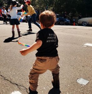 Toddler playing with a rainbow pride flag at an LGBTQIA+ community event, wearing a black North Bay LGBTQI Famililes T-shirt that says Build, Protect, Advocate on the back.