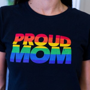 The front of an LGBTQIA+ parent's black t-shirt that says Proud Mom in rainbow striped text.