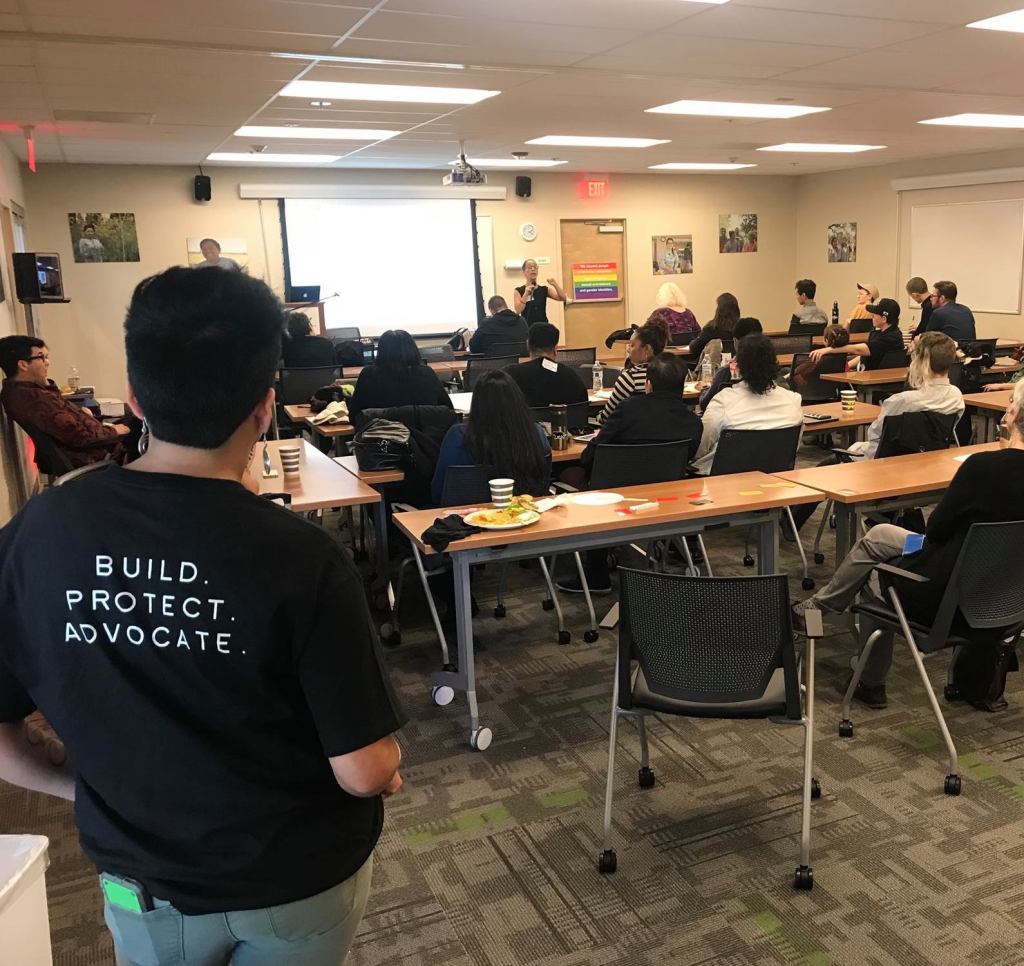 In the foreground, a person in a large room with their back turned to the camera, standing in the back of a room full of tables and people sitting at those tables watching a presentation at a symposium.
